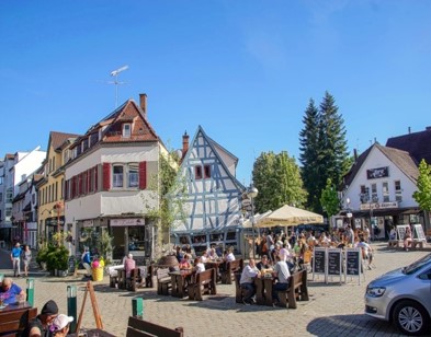 Platz mit Fachwerkhäusern, Menschen, Tischen, Stühen, Schirmen. 