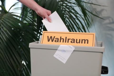 Bild einer Wahlurne mit orangenem Schild mit der Aufschrift Wahlraum, eine linke Hand wirft einen weißen Zettel in die Urne