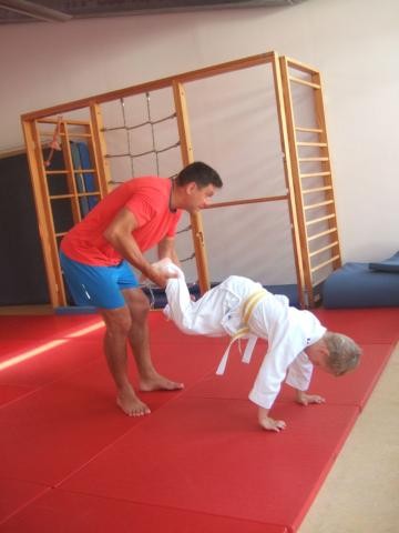 Der Judotrainer hält ein Kind an den Beinen fest. Das Kind berührt mit den Händen den Boden