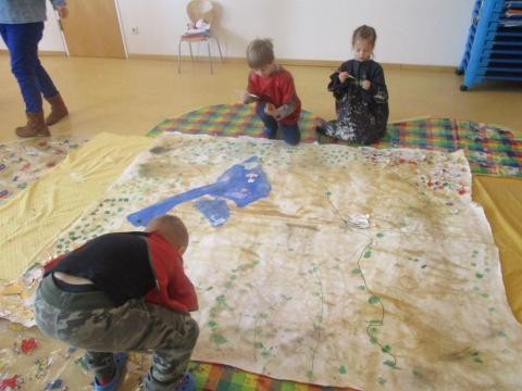 Auf dem Boden liegt ein großes Stück Papier das von drei Kindern mit Farbe bemalt wird