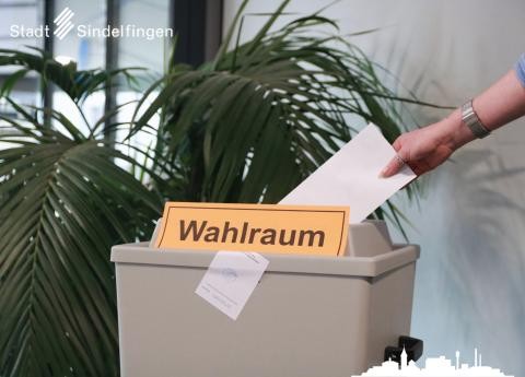 Im Vordergrund mittig eine graue Wahlurne mit orangenem Zettel mit der Aufschrift "Wahlraum" darauf, eine rechte Hand wirft einen weißen Zettel in die Urne. Im linken Hintergrund ist eine grüne Pflanze zu erkennen.